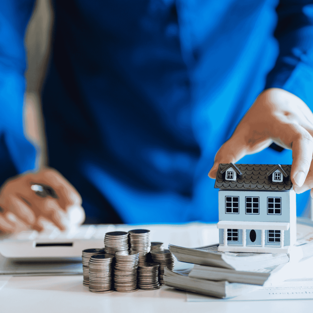 Person in blue shirt handling a house model next to stacks of coins and cash, with a calculator on the table, symbolizing home pricing or real estate finances.