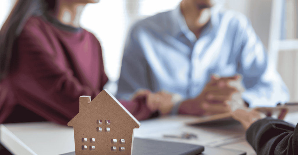 Close-up of a small wooden house model on a desk with two people shaking hands in the background, symbolizing real estate and home buying.