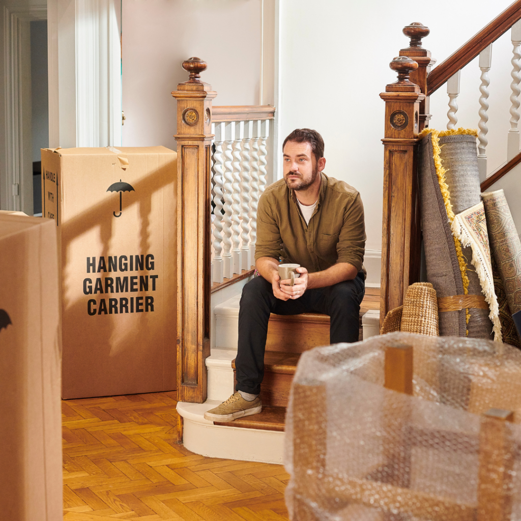 Man sitting on a staircase holding a coffee mug, surrounded by moving boxes and rolled-up rugs inside a home.