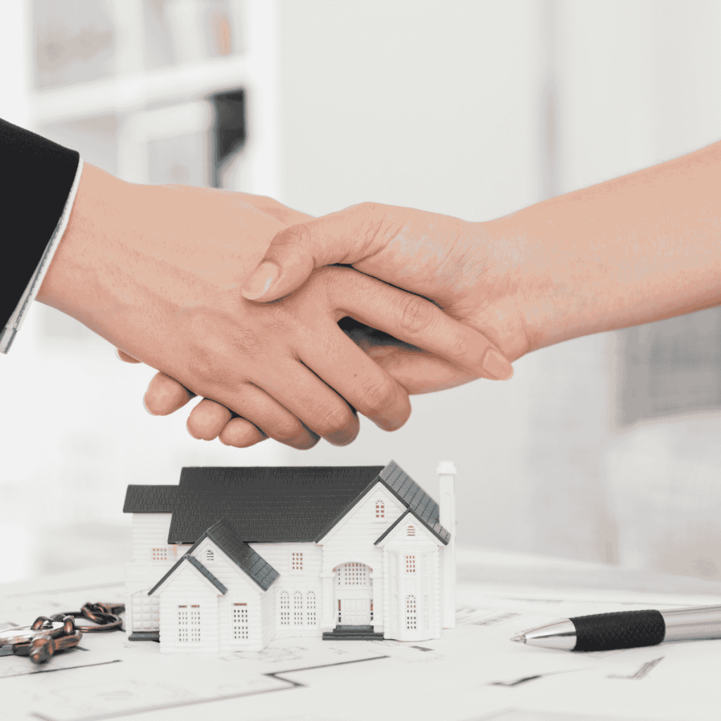 Two people shaking hands over house plans with a model home and keys on the table, symbolizing a real estate deal or home sale agreement.