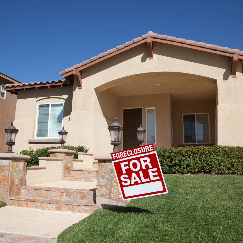 Beige stucco house with a front porch and steps, showing a red 'Foreclosure For Sale' sign on the lawn.