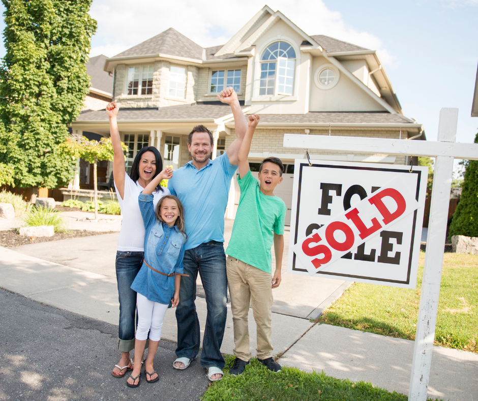 A happy family of four celebrates in front of their new home with a large ‘For Sale’ sign marked ‘SOLD,’ raising their arms in excitement.