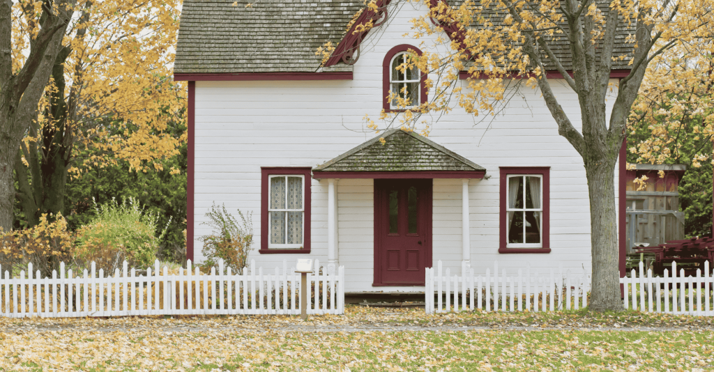 Charming white house with red trim and front door, surrounded by autumn trees and a white picket fence.