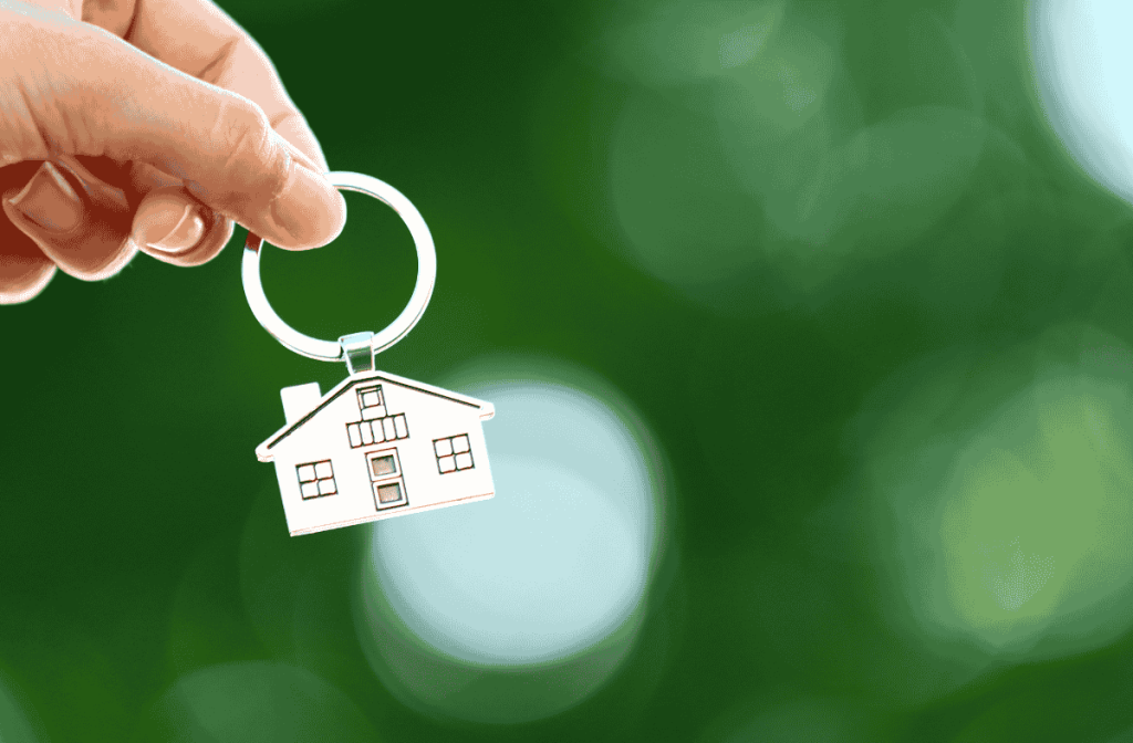 Hand holding a house-shaped keychain against a blurred green background, symbolizing homeownership or real estate.