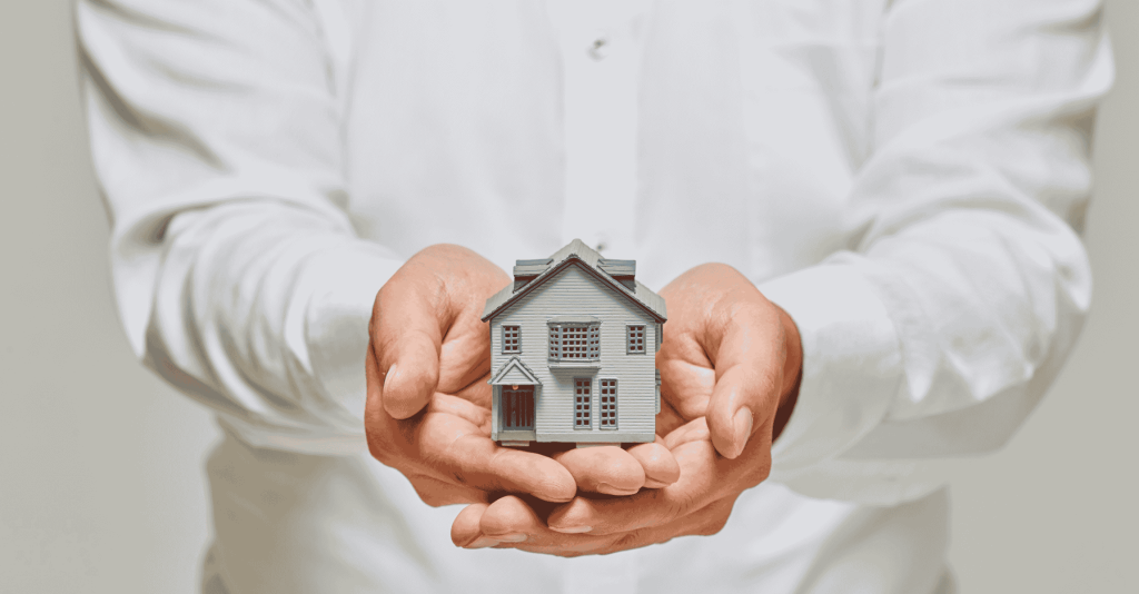 A person wearing a white shirt holds a small model house in their hands, symbolizing real estate, homeownership, or property investment.
