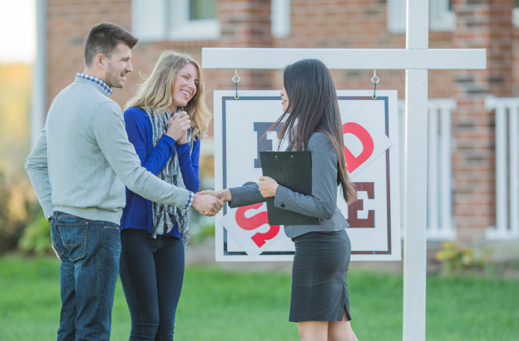 Smiling couple shaking hands with a real estate agent in front of a home with a For Sale sign.
