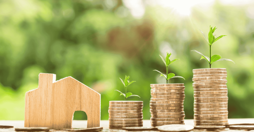 Stacks of coins with small plants growing on top next to a wooden house model, symbolizing real estate investment growth.