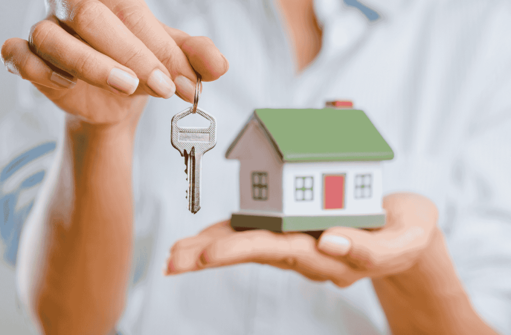 Person holding a house model in one hand and a key in the other, symbolizing home ownership or real estate sales.