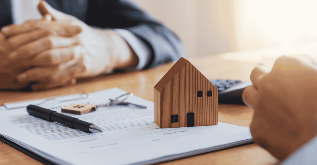 Real estate agent and client discussing a home sale contract with a small wooden house model, pen, and keys on the table.