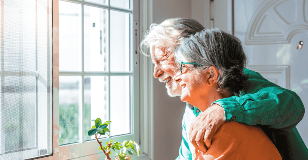 Smiling senior couple embracing while looking out a bright window inside their home.