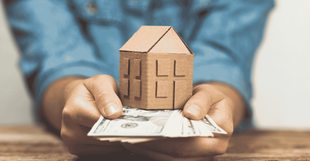 Person holding a cardboard model house and a stack of U.S. dollar bills, symbolizing real estate investment and home value.