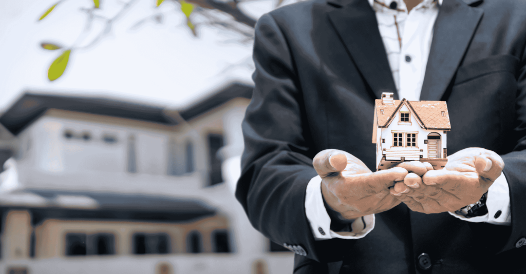 Real estate agent in a suit holding a small model house in his hands in front of a large home.