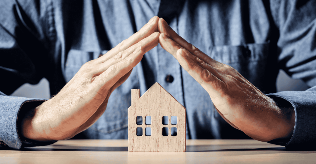 Person forming a protective roof shape with their hands over a small wooden house model, symbolizing home protection and organization.