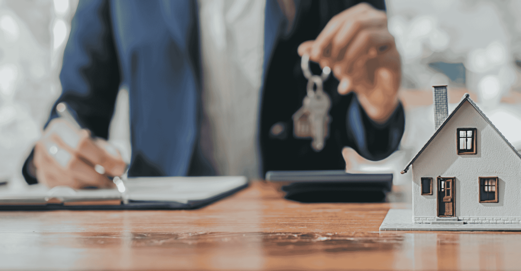 Person signing real estate documents while holding house keys, with a small model house on the table representing a property sale or closing.