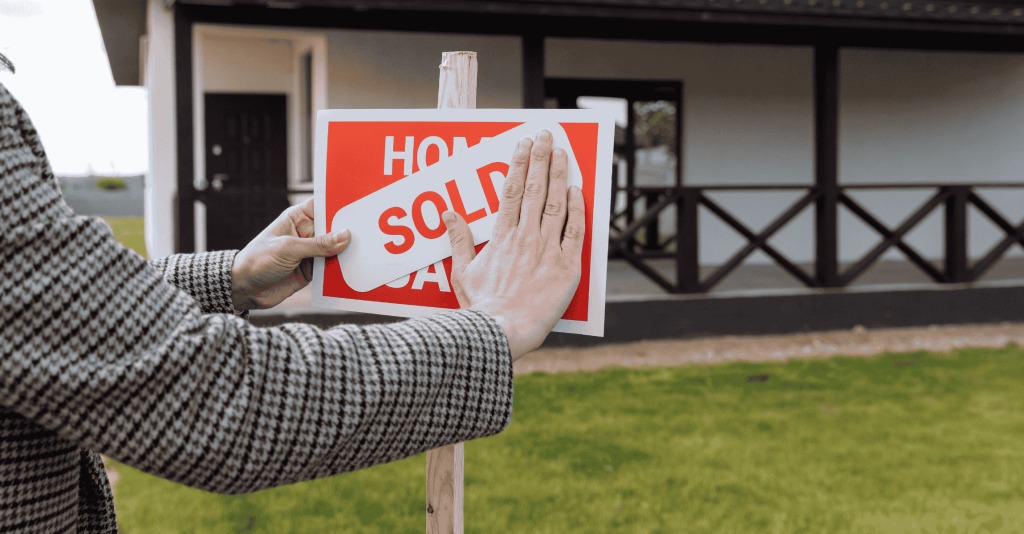 Person placing a “SOLD” sticker over a “For Sale” sign in front of a house, symbolizing a successful home sale.
